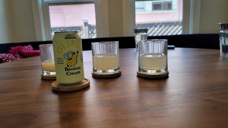 Photo of can of OLIPOP Banana Cream soda with a yellow cartoon minion waving from it. Behind are three glasses with various levels of a hazy, lemonade-colored liquid