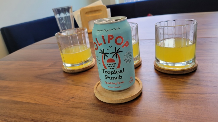 Photo of can of OLIPOP Tropical Punch sparkling tonic in a sea blue can with stylized palm trees. Behind the car are three glasses with a yellow liquid