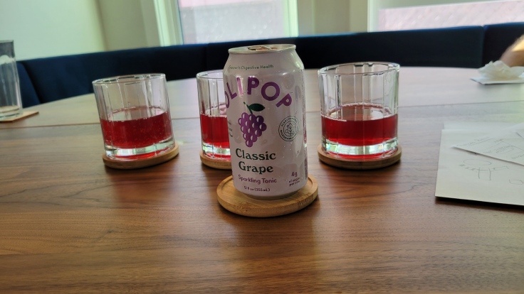 Photo of can of OLIPOP Classic Grape sparkling tonic in a light purple can with a stylized cluster or purple grapes. Behind the car are three glasses with a ruby red liquid